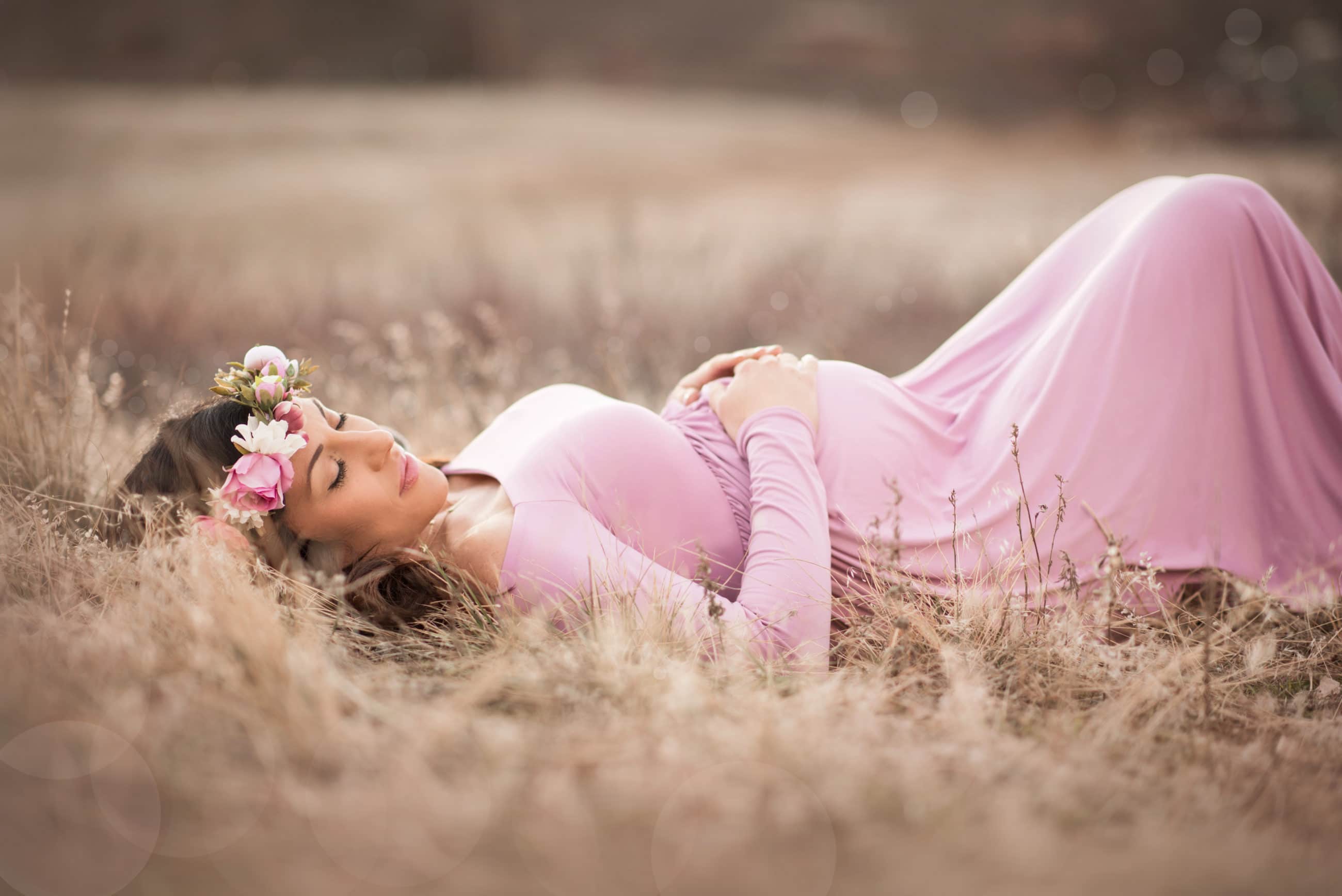 pregnant mother in pink maternity gown and flower crown laying down in golden grass - baby's first year - boulder newborn photographer
