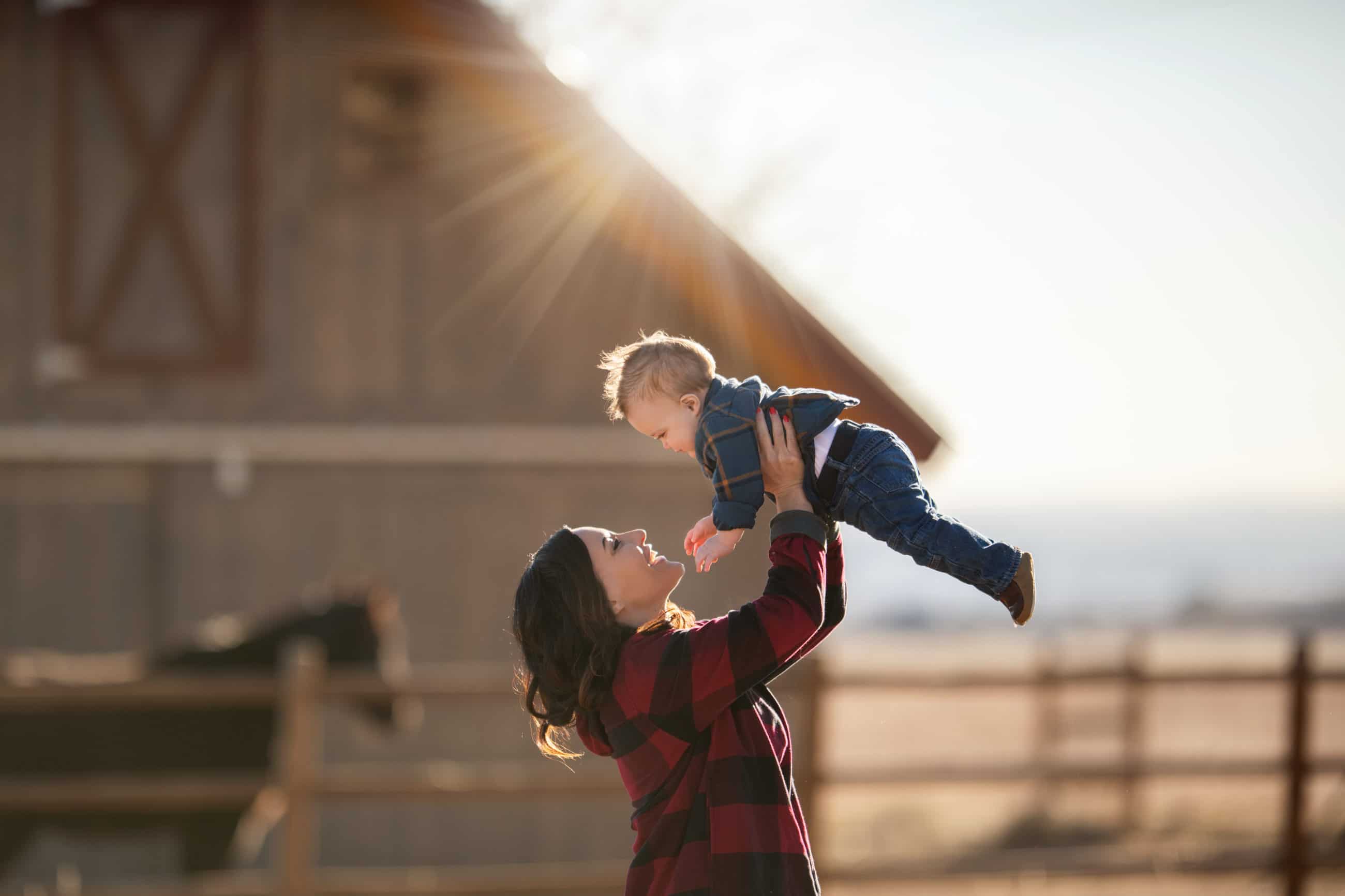 mom holding baby boy up into the air barn and black horse in background - boulder family photographer