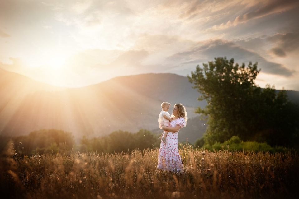 mom holding baby girl in golden light in field of gold with mountains in Colorado Mother daughter photo session | boulder family photographer